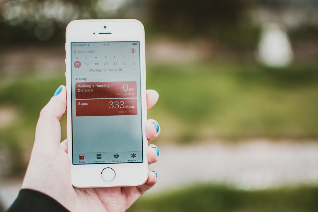 A person holds a smartphone showing a fitness app with step count and distance tracking outdoors.
